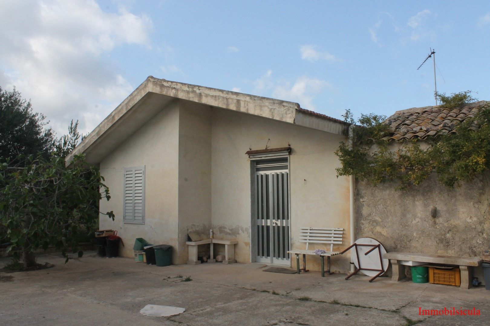 Piccola casa beige con tetto spiovente, porta d'ingresso e mobili da esterno sparsi in un cortile sotto un cielo azzurro.
