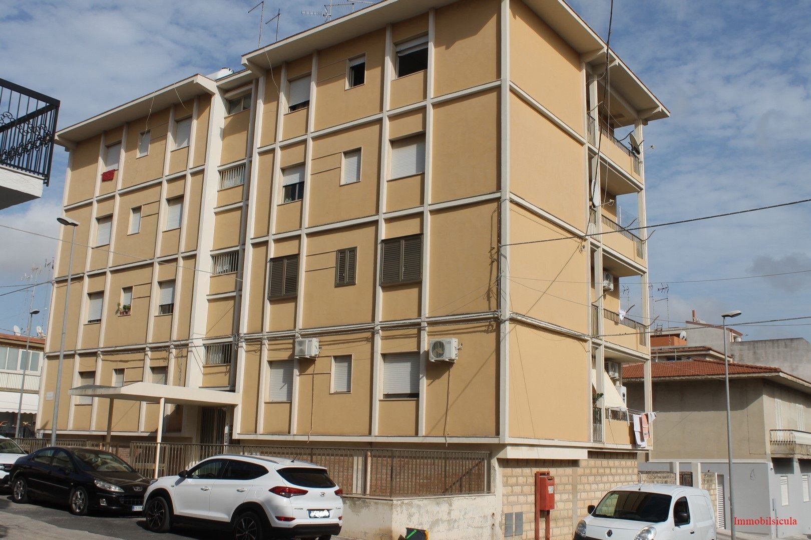 Palazzo di appartamenti beige con balconi e auto parcheggiate su una strada sotto un cielo nuvoloso.