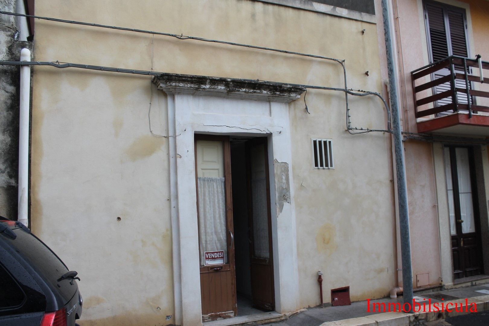 Facciata di un piccolo edificio beige con una porta stretta, una finestra con sbarre e un balcone sulla destra.