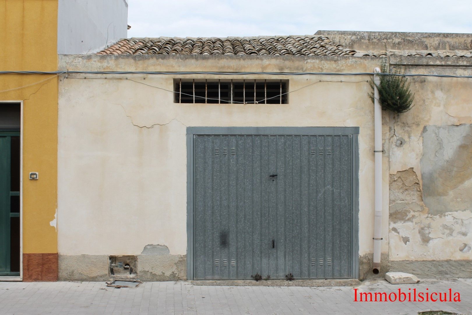 Piccolo edificio beige con una porta da garage in metallo grigio e una stretta finestra in alto, accanto a un muro giallo.