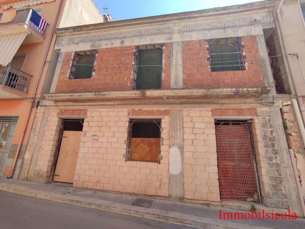 Edificio a due piani in mattoni non rifiniti con finestre murate, situato su una strada in pendenza.