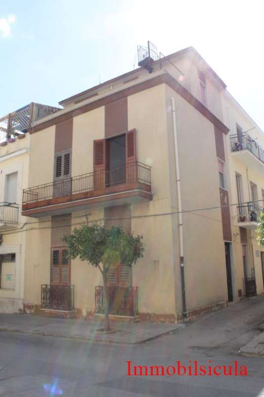 Edificio d'angolo a due piani di colore beige con persiane marroni, balcone e un piccolo albero, situato in una strada stretta.