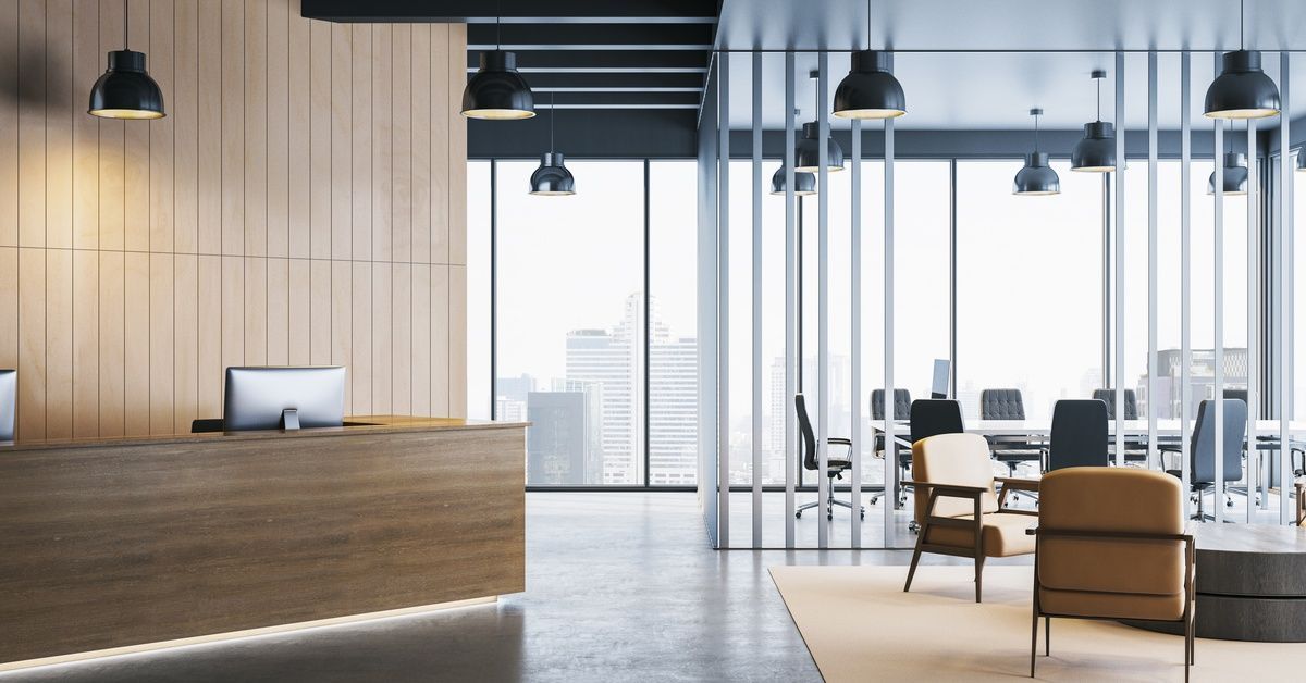 An office lobby with a desk and computer to the left of frame and several chairs and windows to the right.