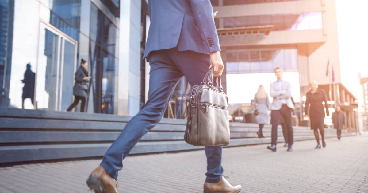 Closeup of a man’s lower body dressed in a suit and holding a brief case as he walks outside an office building.