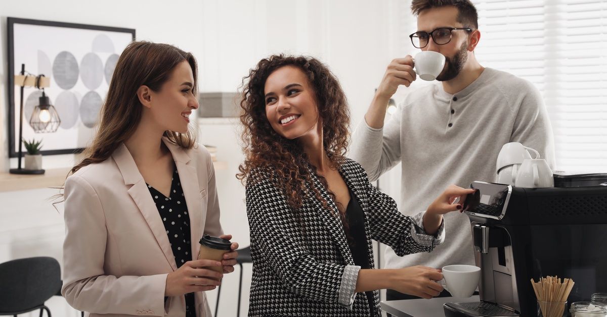 Three young business professionals standing and talking in an office break room and kitchen while getting coffee.
