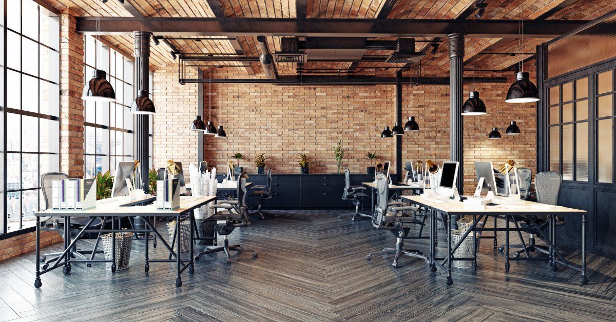 A large, open office space with several desks, gray wooden floors, brick walls, and floor to ceiling windows along one wall.