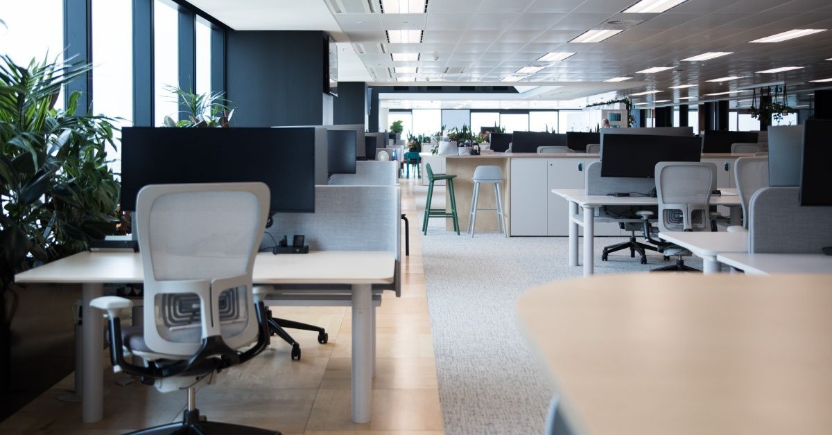 A large room with many desks; each desk has a PC monitor on it. There are windows lining the walls.