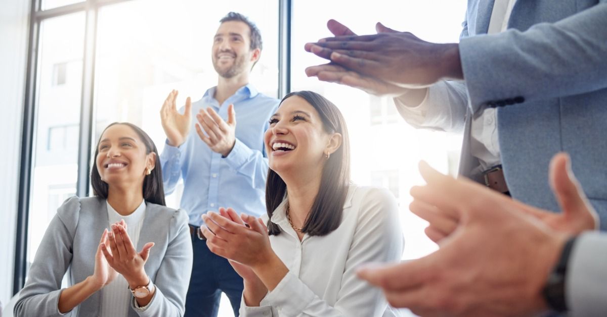 A group of people in business clothes are all smiling and clapping. Some are sitting in chairs while others are standing.