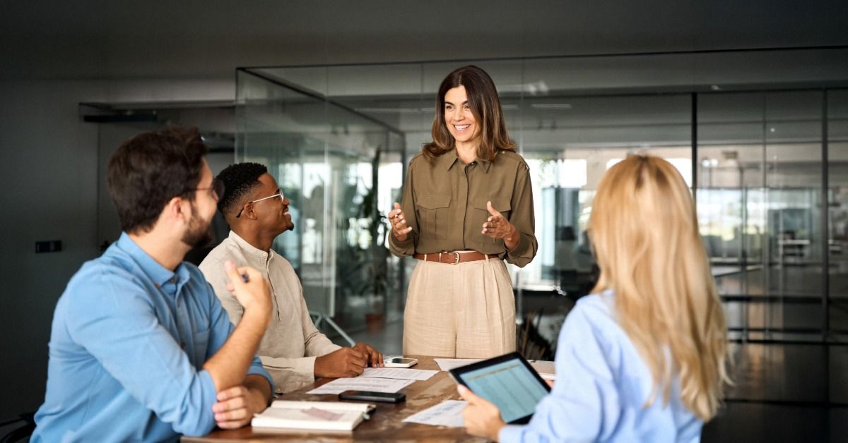 A woman is standing at the head of a table as others, who are sitting down, look up to her.