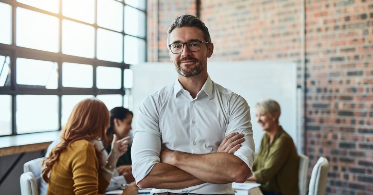 A man with folded arms is leaning back against a table. Sat at the table are several people.