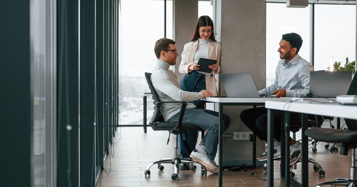 Two men are sitting at a table while a woman with a tablet in her hands is standing next to them.