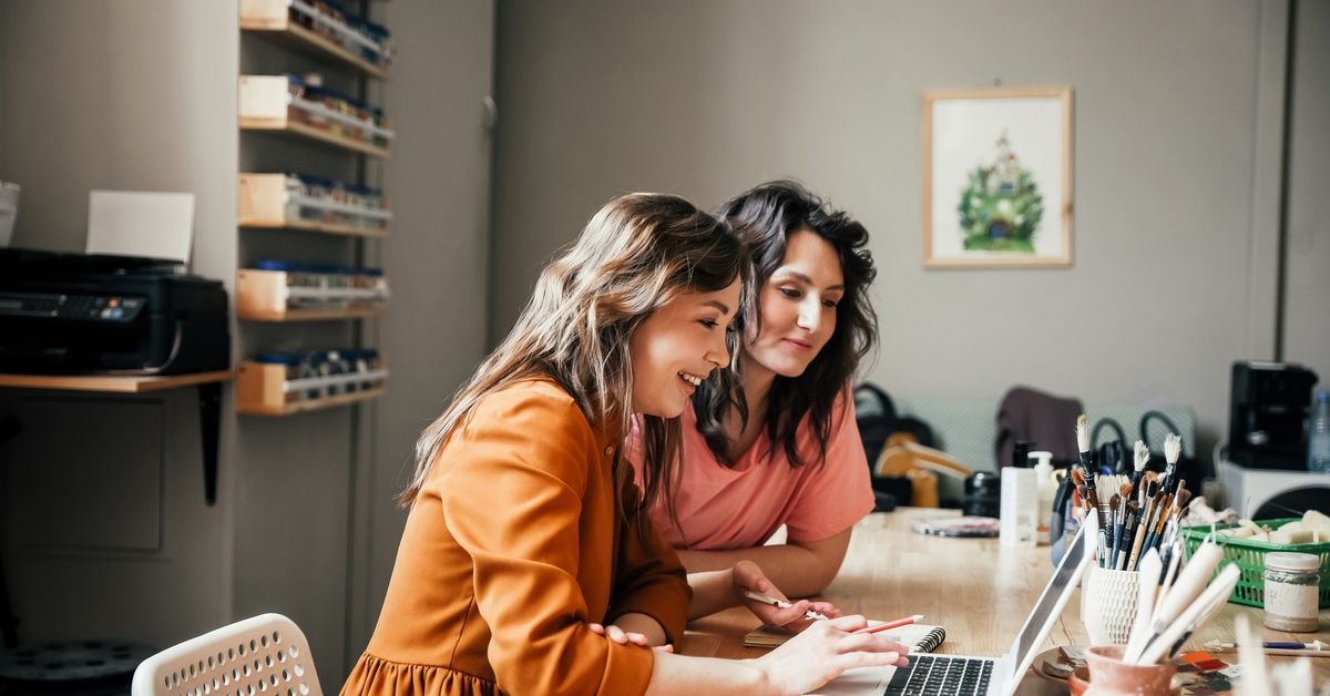 Two women are sitting at a desk. One woman is using a laptop while the second woman is watching.