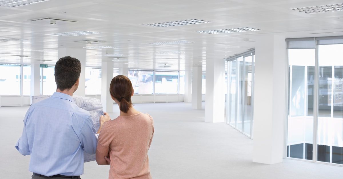 A man and a woman are speaking while looking at a bright, empty office. The man holds up a sheet of blueprints.