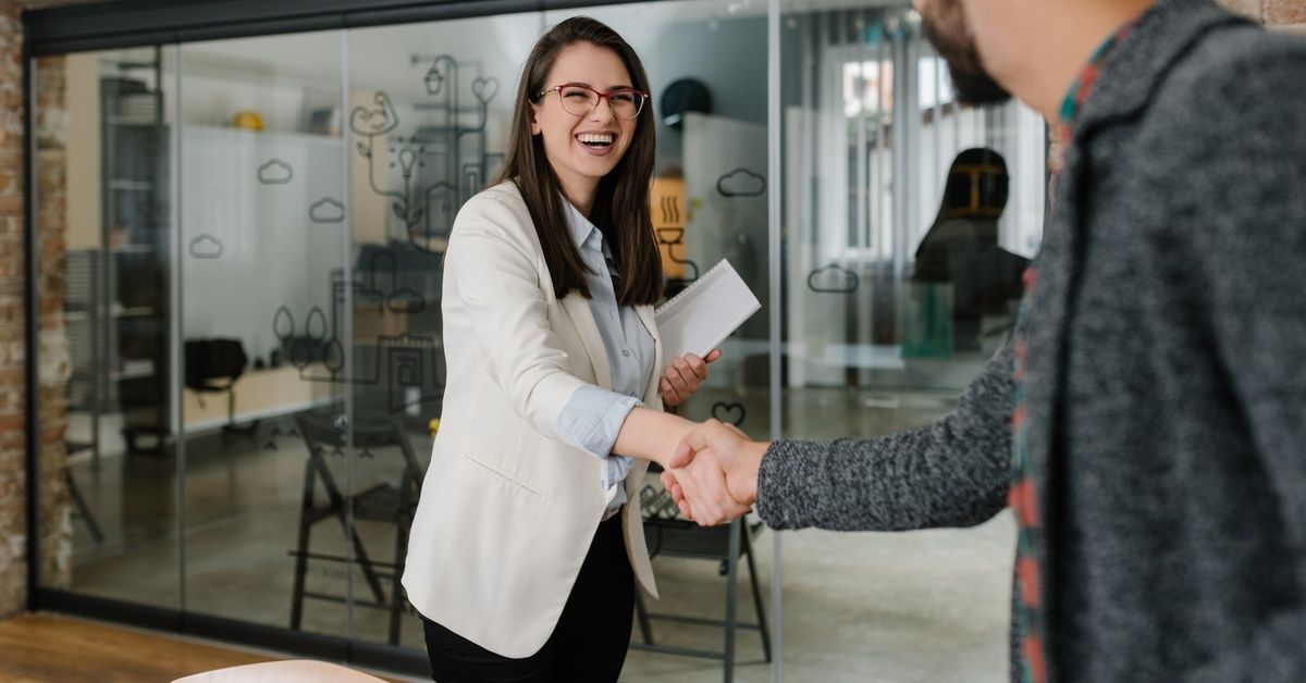 A man and woman shake hands inside an office space with glass walls, hardwood floors, and brick accents.