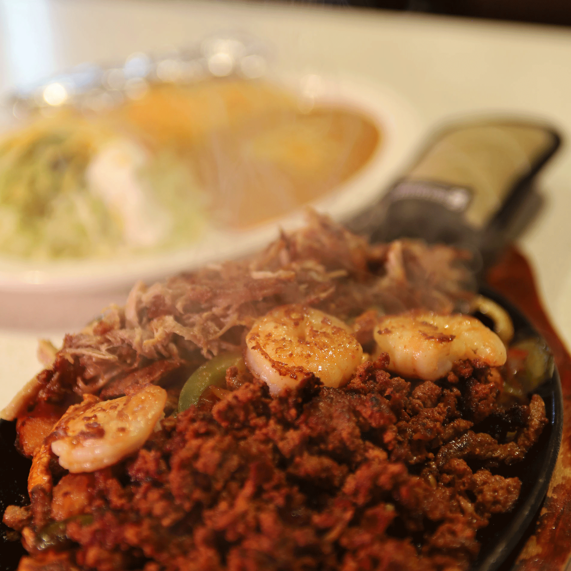 Sizzling fajitas with shrimp, chorizo, and shredded meat on a wooden board. Blurred burrito in background.