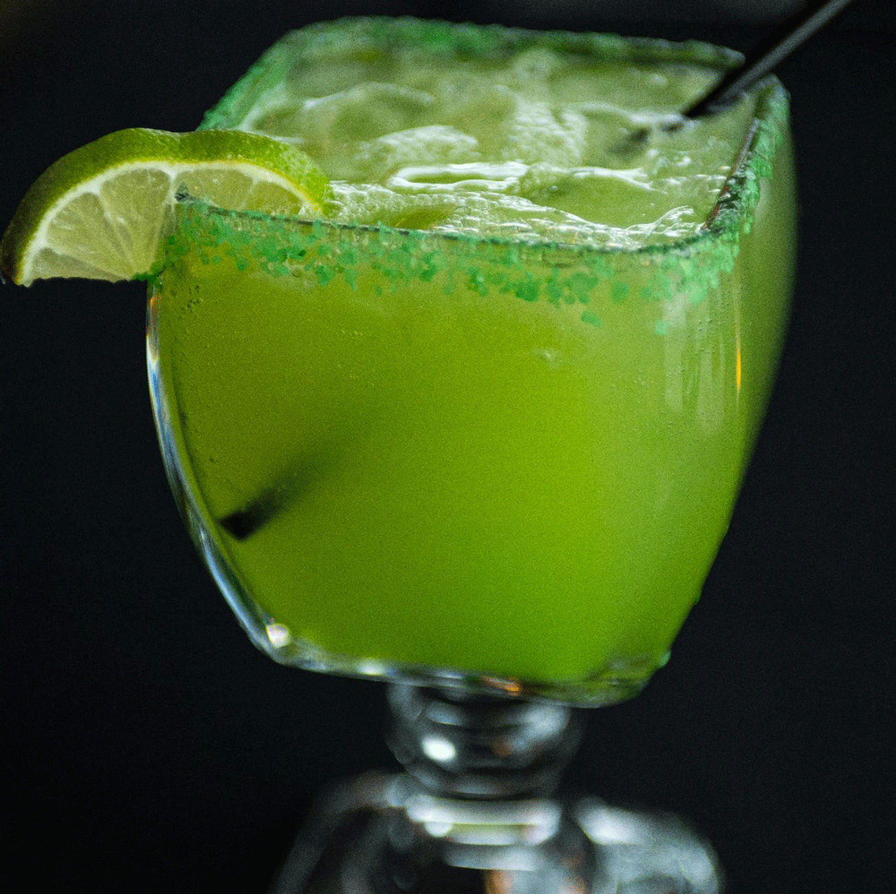 Green margarita in a square glass with lime wedge and green sugar rim.