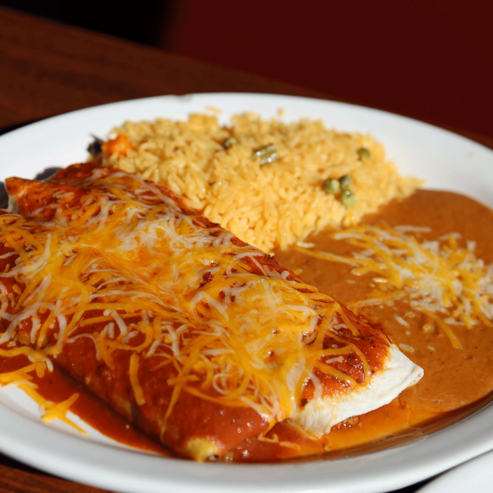 Enchiladas covered in melted cheese, served with rice and refried beans on a white plate.