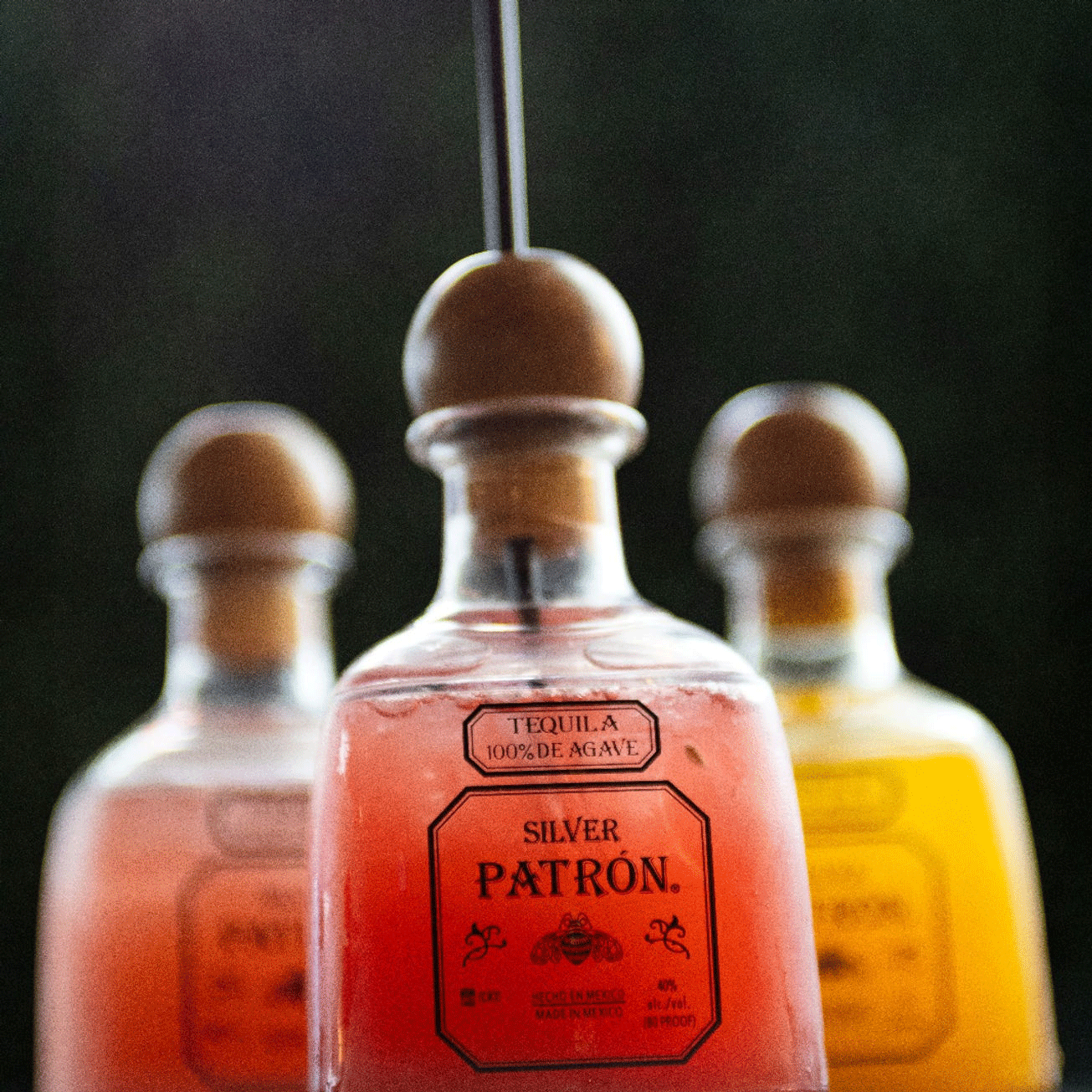 Three Patron tequila bottles with colorful liquids and cork stoppers. A straw is in the center bottle.