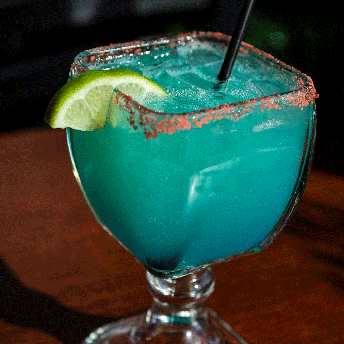 Blue cocktail in a square glass, rimmed with red salt, lime wedge garnish.