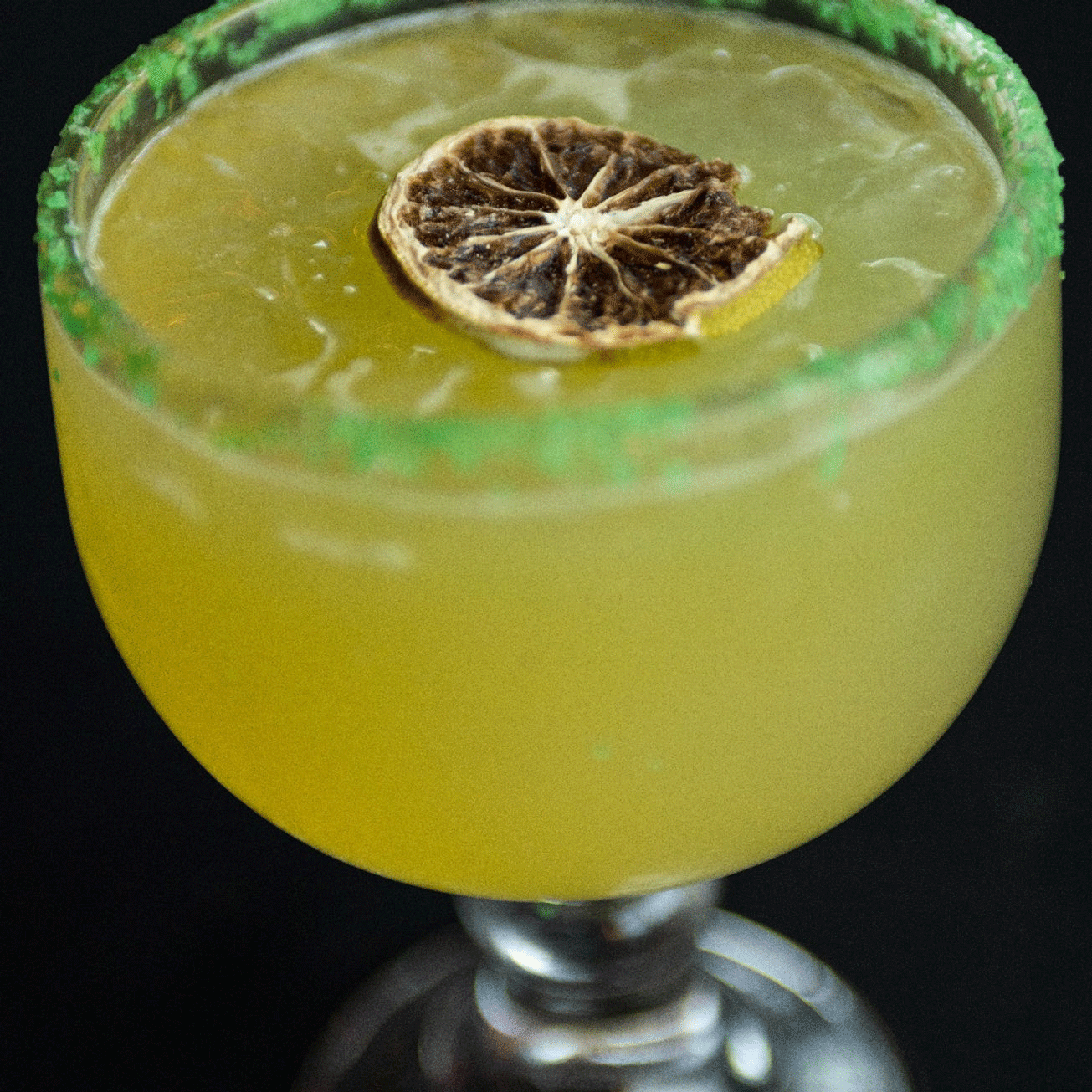 Yellow cocktail in a stemmed glass, green sugar rim, dried lime garnish.