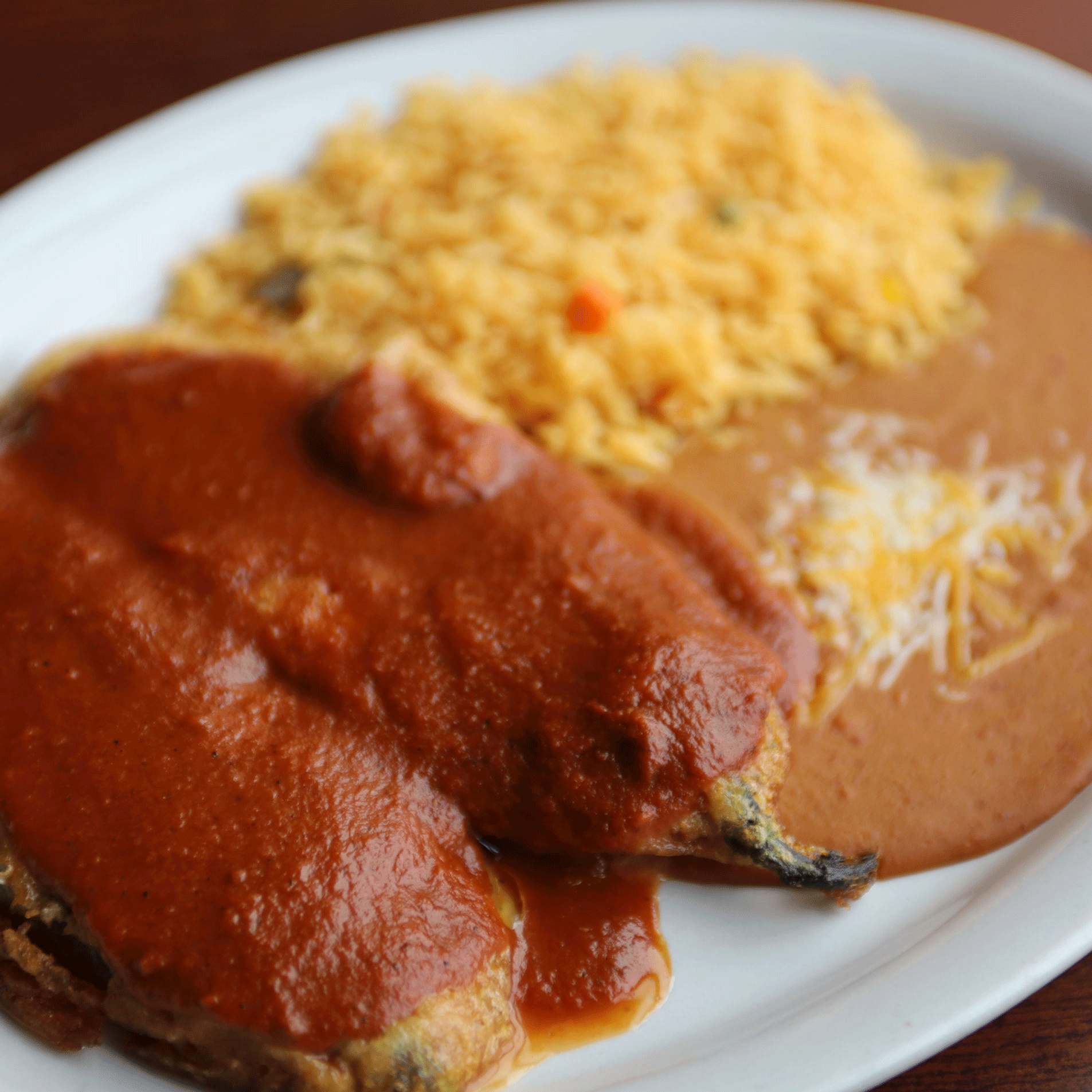 Chile relleno with red sauce, rice, and refried beans on a white plate.