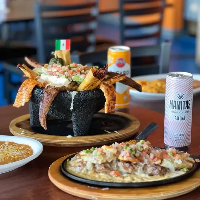 Mexican food spread: nachos in a molcajete, sizzling skillet with nachos, canned drinks, and a side of beans.