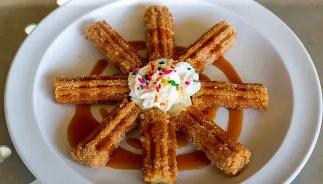 Churros arranged on a plate with whipped cream, sprinkles, and caramel sauce.