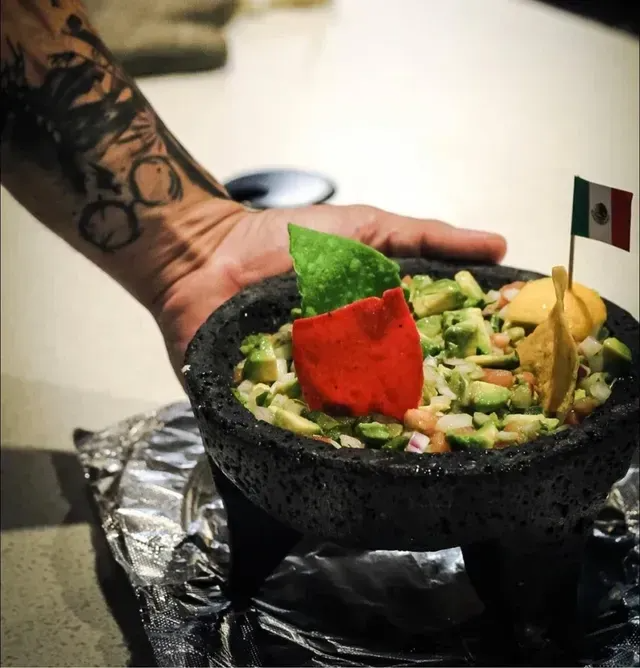 Hand holding a bowl of guacamole with chips and a Mexican flag on a table.