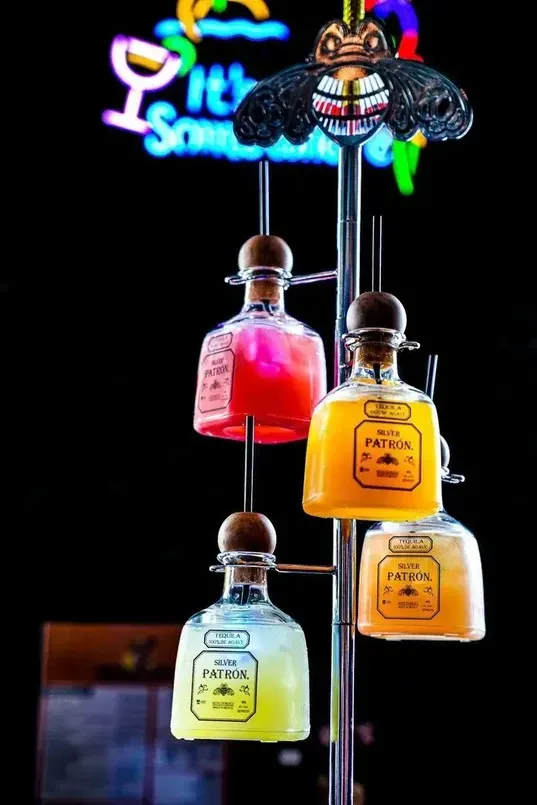 Tequila cocktails in Patron bottles hanging from a pole with a tiki head and neon sign.