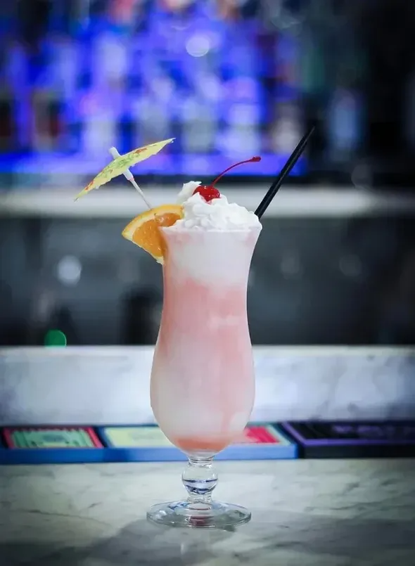 Pink cocktail in a tall glass, garnished with whipped cream, orange slice, cherry, and umbrella.