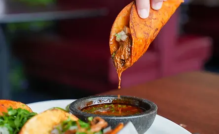 A person dips a taco filled with meat into a bowl of sauce; plate of tacos in the foreground.