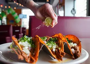 Person squeezing lime juice over tacos on a plate in a restaurant.