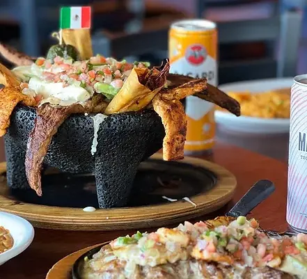 A large volcanic rock bowl overflowing with Mexican food, drinks, and a small flag on a table.
