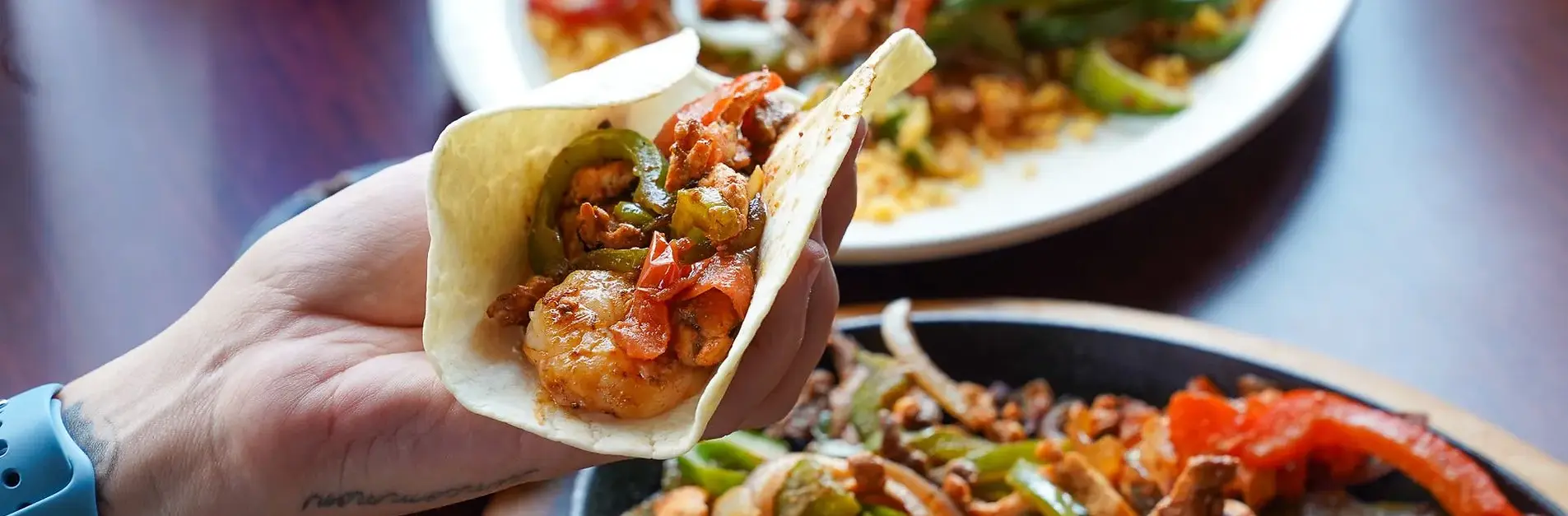 Hand holding a taco filled with shrimp and vegetables, with a plate of food in the background.
