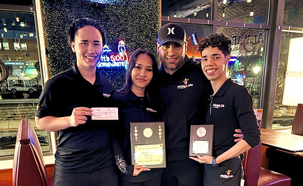 Four people at a restaurant, holding awards and a check. They wear black shirts and pose near a window.