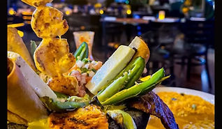 Close-up of a Mexican dish: grilled meat skewer, jalapeños, cheese, and tortilla chips in a bowl. Dim restaurant setting.