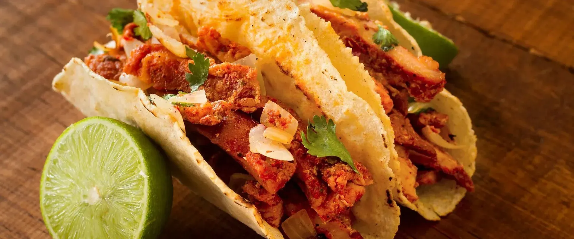 Three tacos with meat filling, cilantro, and lime on a wooden surface.