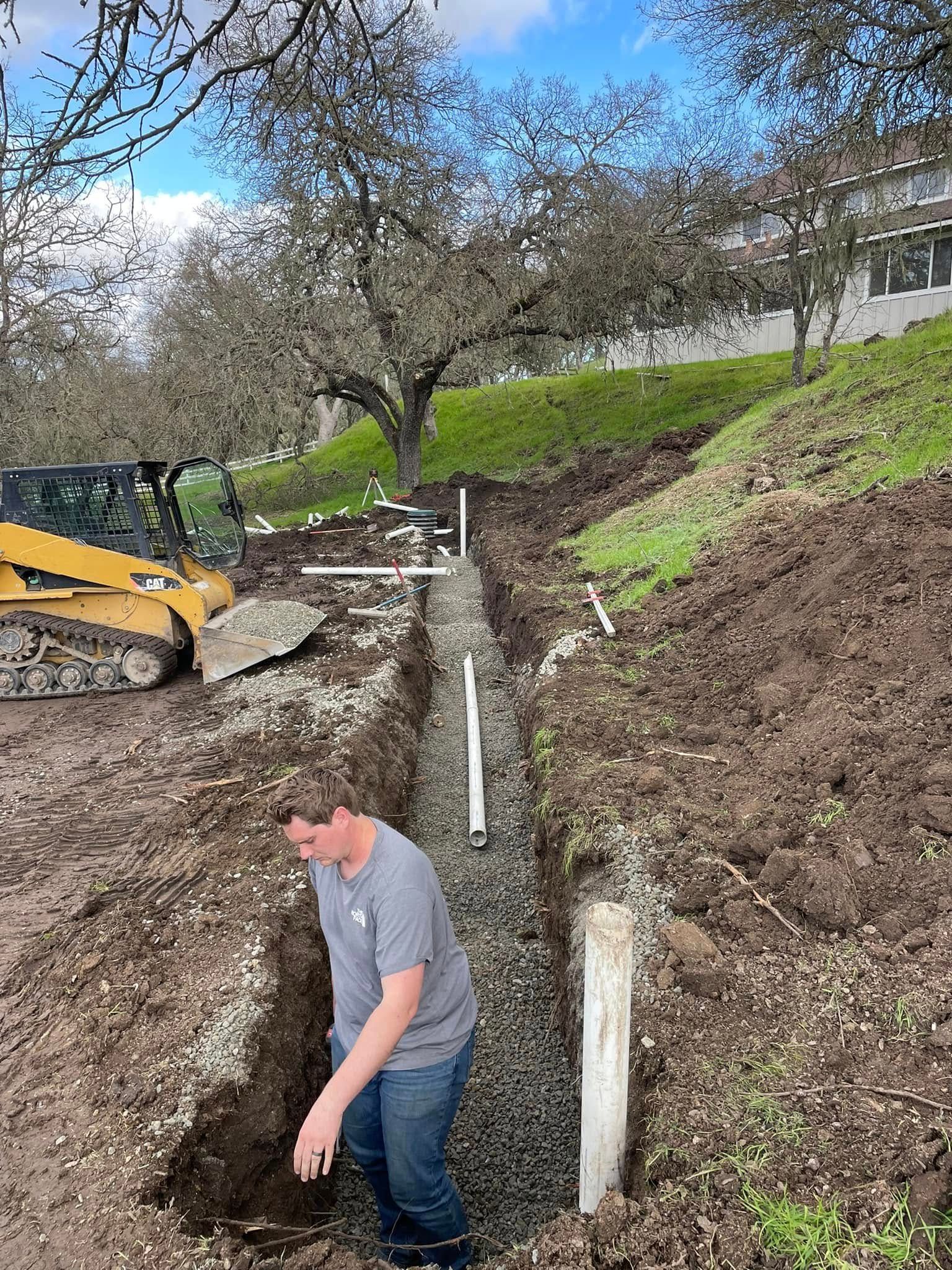 Man digging a trench beside a grassy hillside with an excavator and white posts in the background