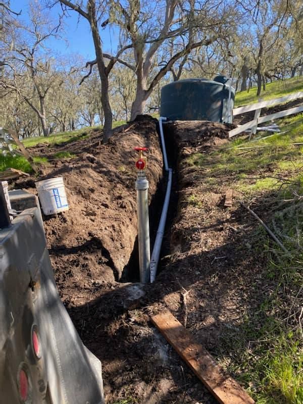 Utility trench with exposed pipes and a large black tank in a grassy, wooded slope