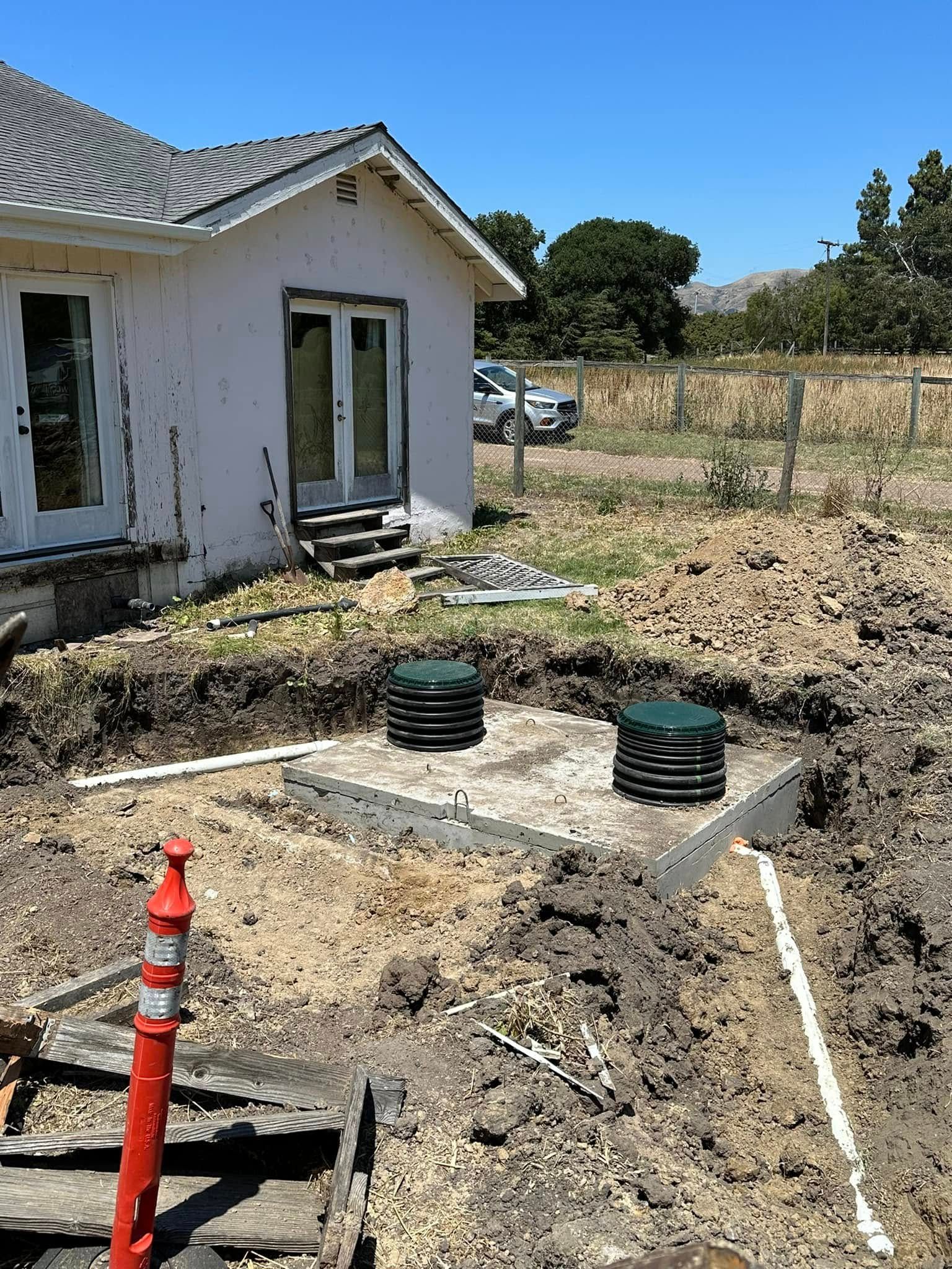 Construction site beside a white house, with a dug trench, pipes, and two green utility covers.