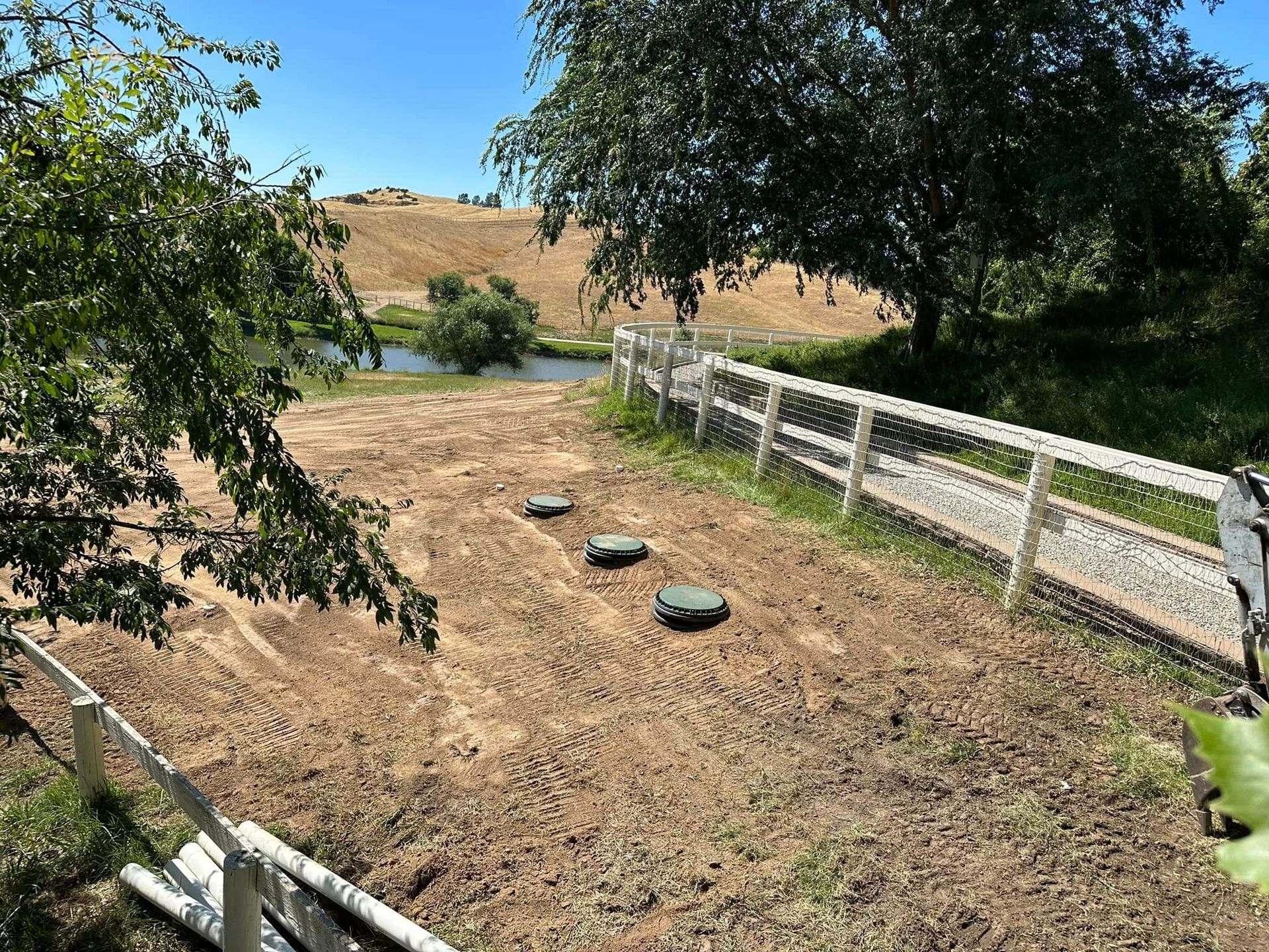 Dry path with stepping stones beside a white fence under trees, leading to a pond and hills beyond.