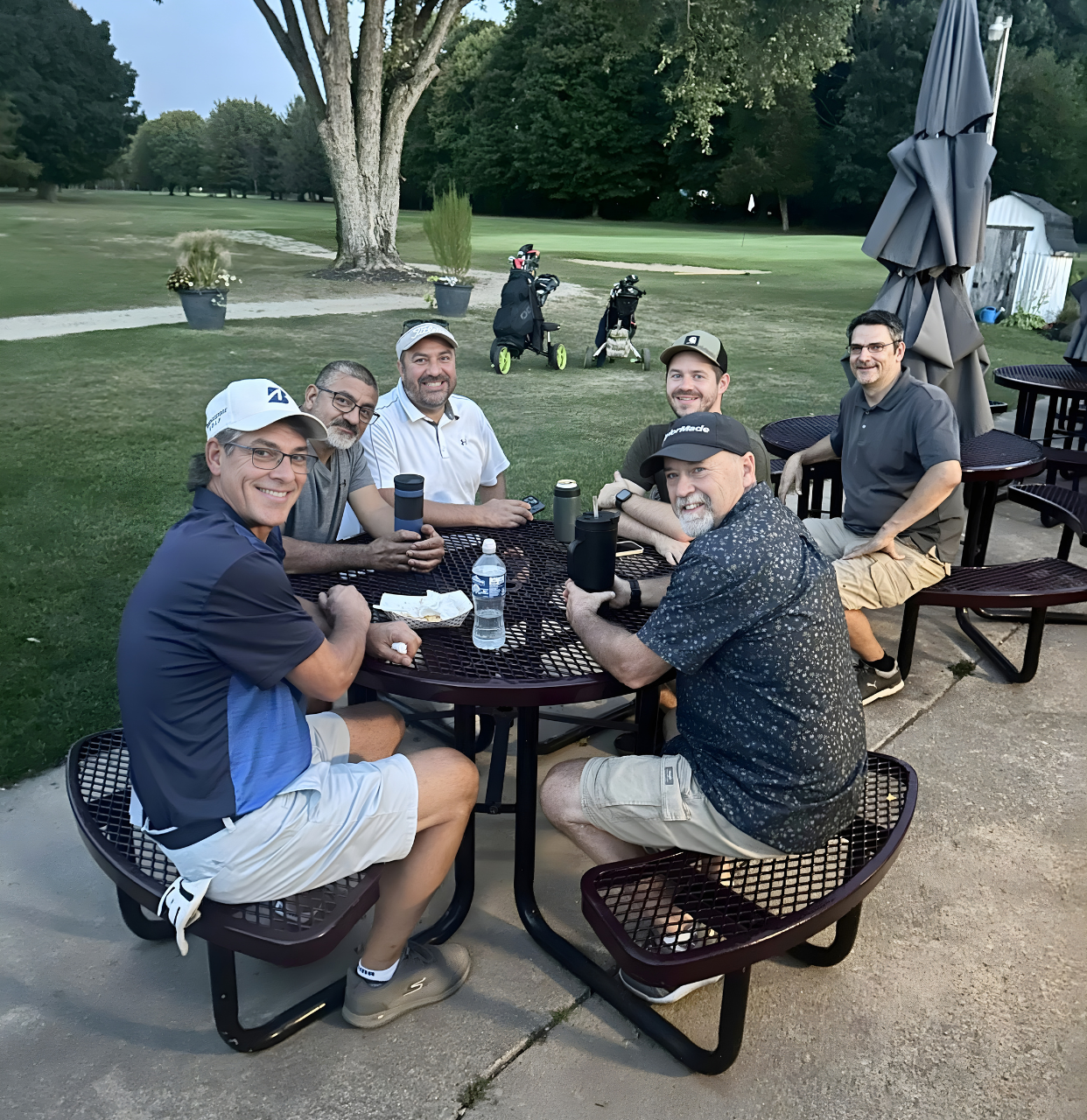 Six people smile around a round outdoor table at a golf course at dusk.