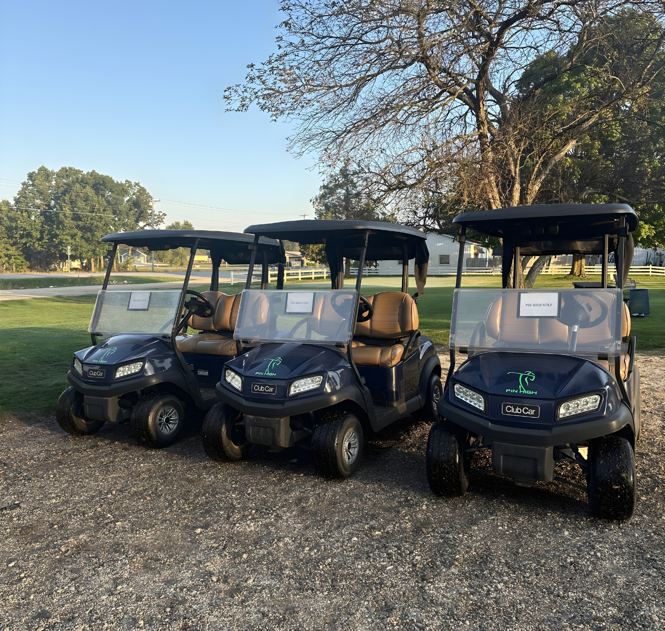 Three dark-colored golf carts with tan seats parked side-by-side on a gravel lot near a grassy area and trees.