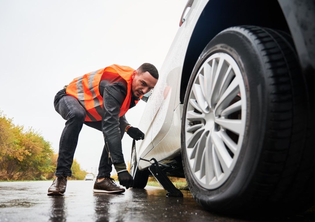 Man in orange vest changing a flat tire on a wet road.