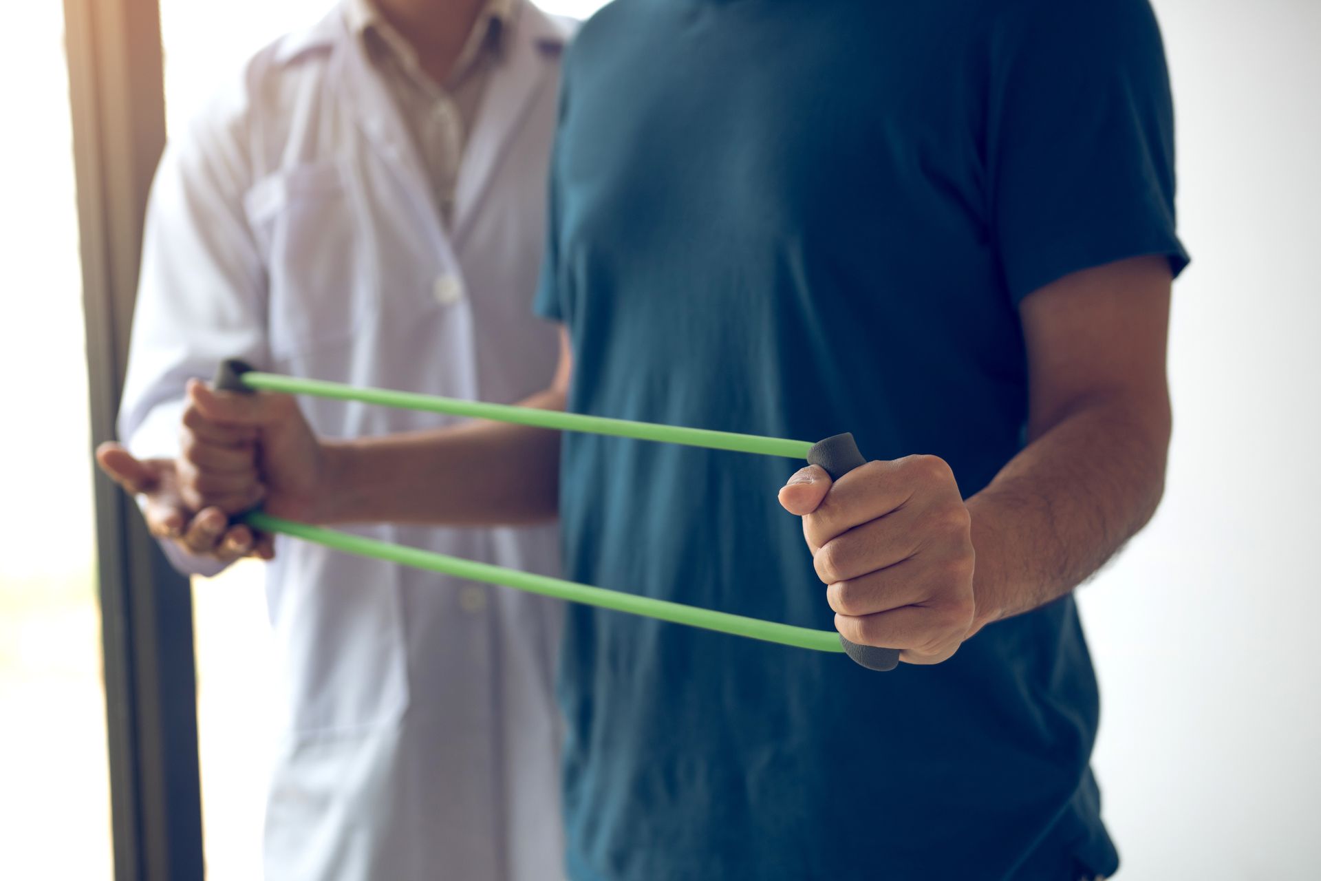 Two people stretching a green resistance band in a bright gym or therapy room.