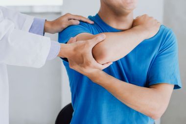 Physical therapist assisting a seated patient with an arm exercise in a bright clinic room