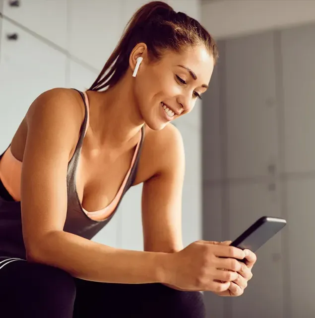 Woman in a sports bra smiling at her phone while leaning over indoors, wearing wireless earbuds