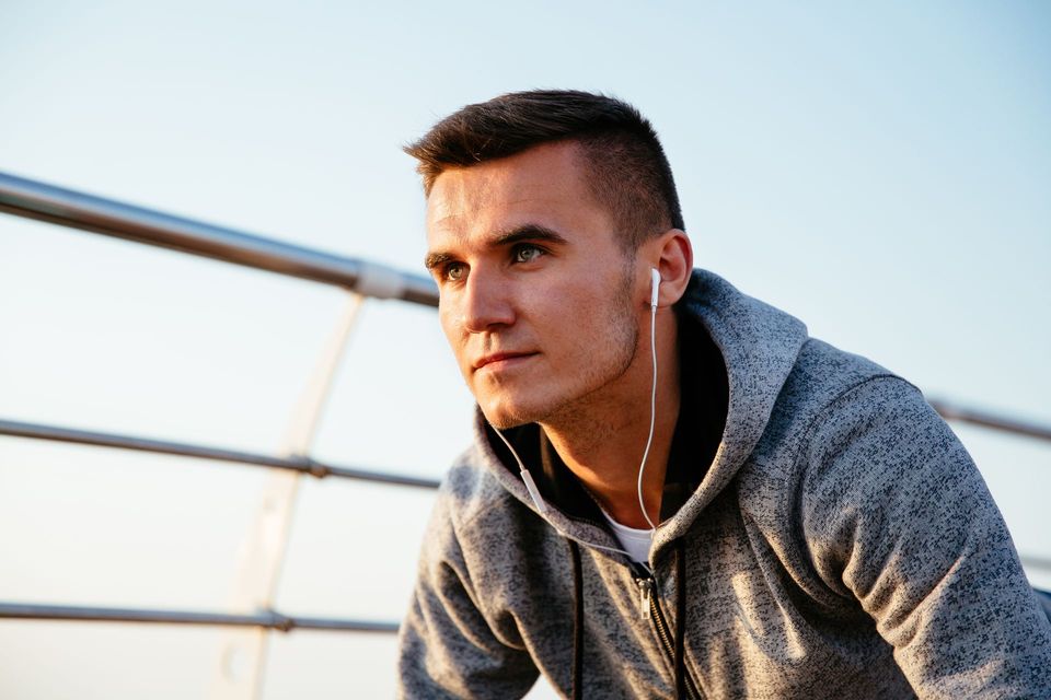 Young man in a gray hoodie leaning forward outdoors with earbuds, looking ahead thoughtfully