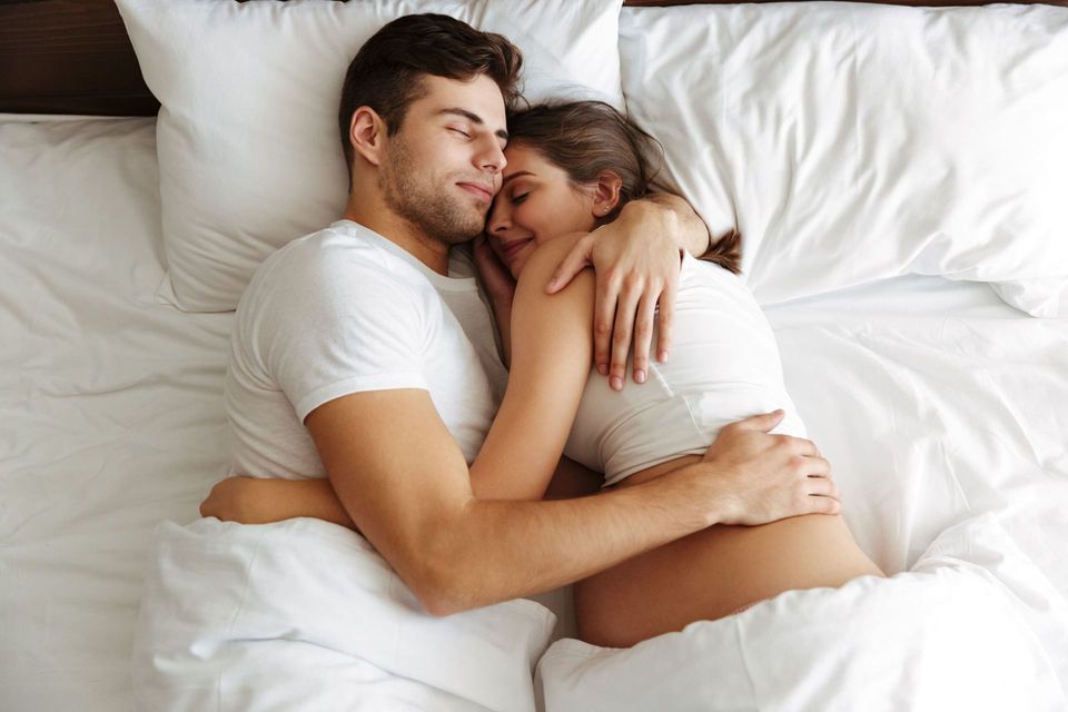 Couple cuddling in bed under white sheets, eyes closed, in a cozy, intimate pose