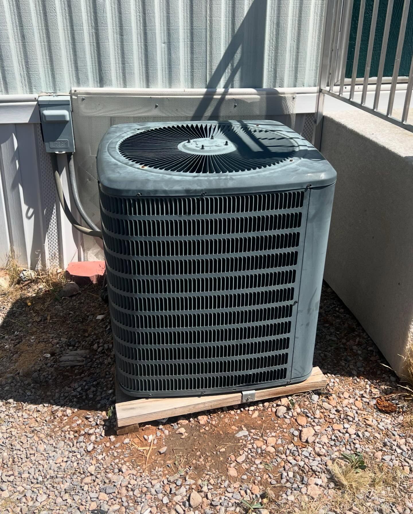 Outdoor central air conditioner unit beside a house wall and railing on gravel